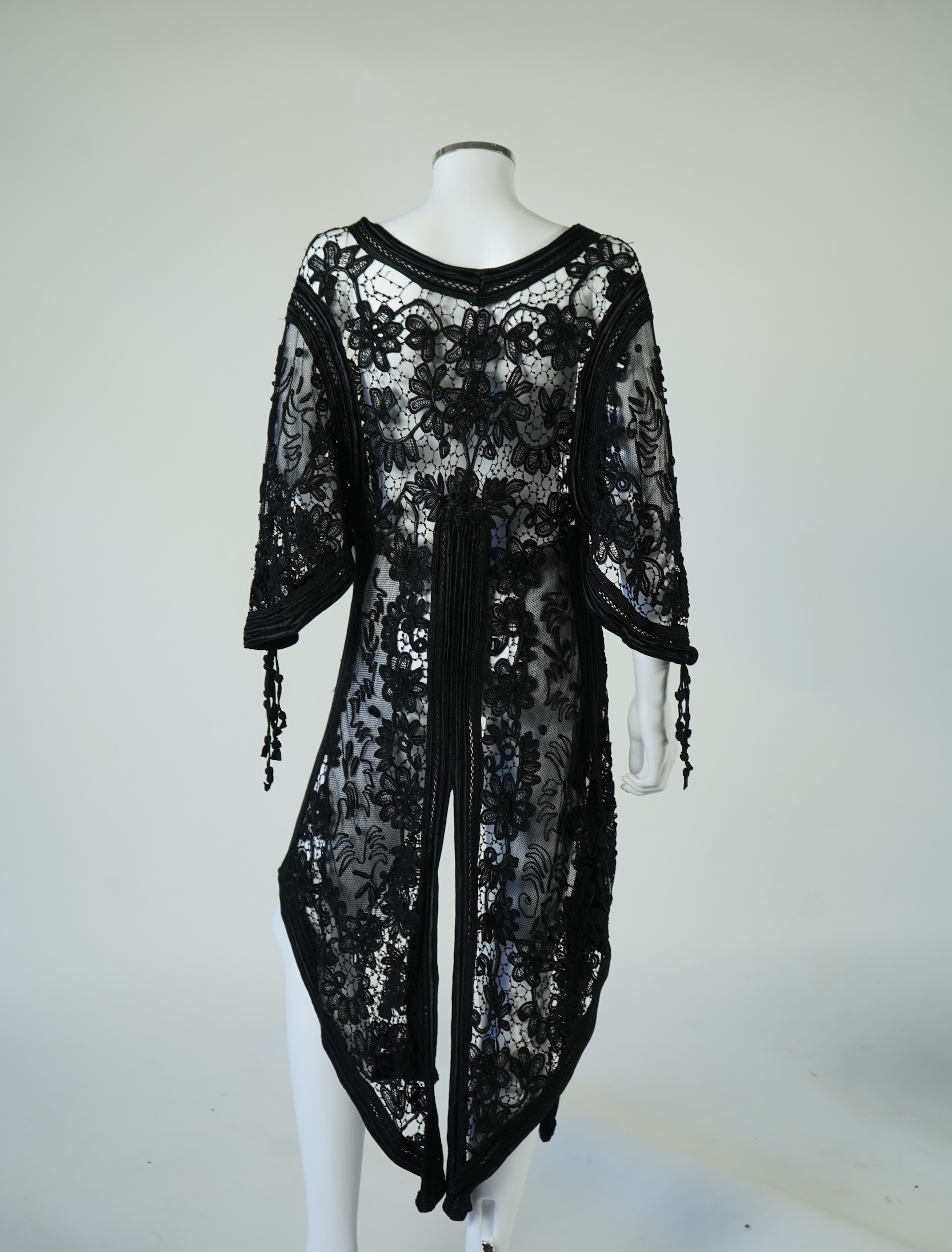 A late 19th century stylish ornate black silk tape lace evening coat/dress. Designed with a wide open neck, short bell sleeves with long tassels, four large buttons at the front and tails at the front and back. 130cm lon